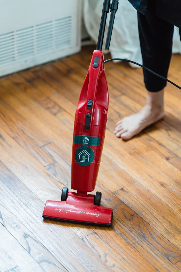 A red vacuum cleaner on a wooden floor with an adults foot visible, indicating home cleaning.